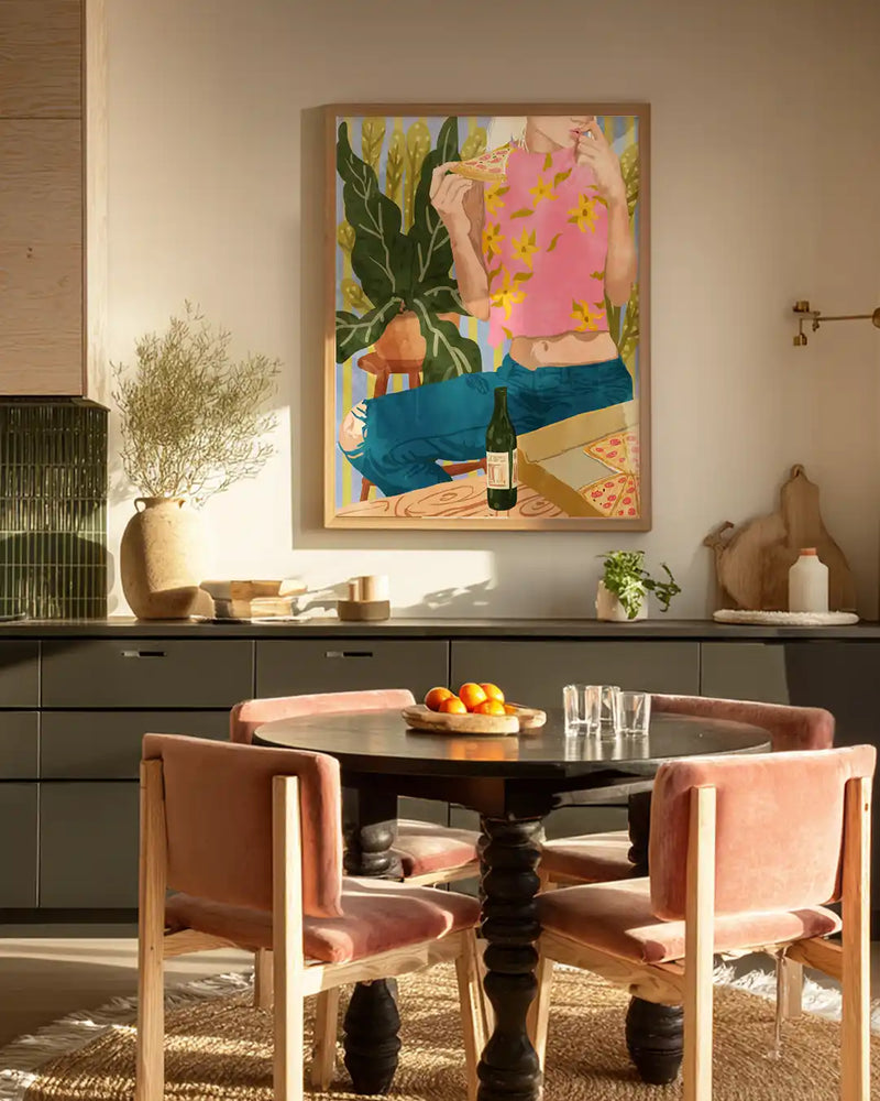 A modern dining room interior featuring a framed contemporary wall art print of a woman enjoying pizza and wine.The artwork adds a playful, artistic touch above a sleek dining table with soft pink upholstered chairs.Neutral cabinetry, natural textures, and warm lighting create a cozy yet stylish atmosphere.Perfect inspiration for modern home decor, dining room wall art, and contemporary interior styling.