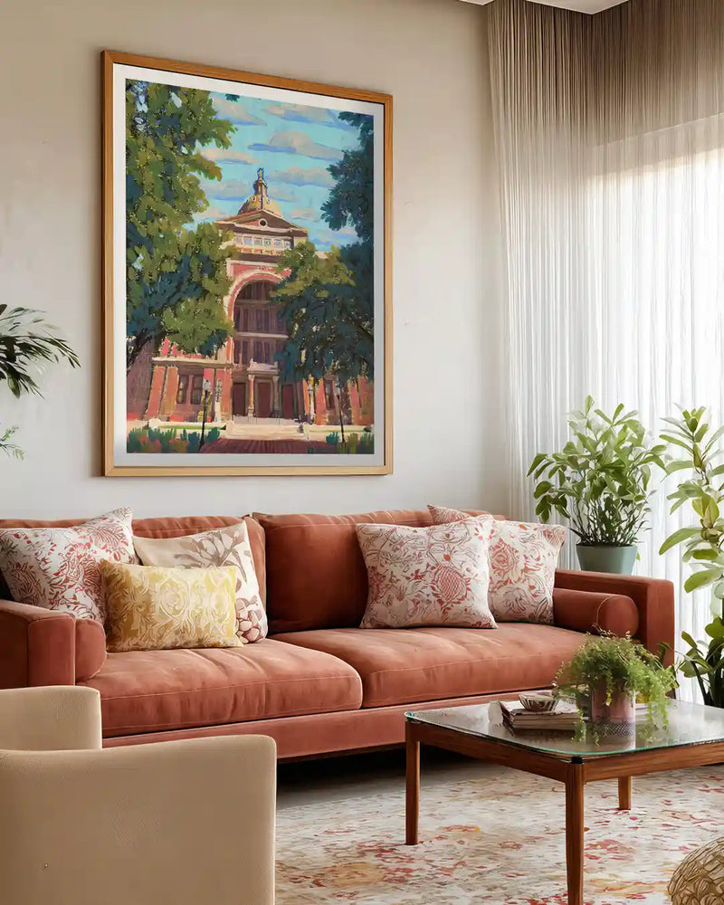 A bright contemporary living room featuring a large framed architectural wall art print above a rust-colored sofa.The artwork showcases a classic building surrounded by lush trees, adding a sophisticated artistic touch.Neutral tones, patterned cushions, indoor plants, and sheer curtains create a warm and elegant interior style.Perfect inspiration for modern home decor, living room wall art, and stylish interior design ideas.