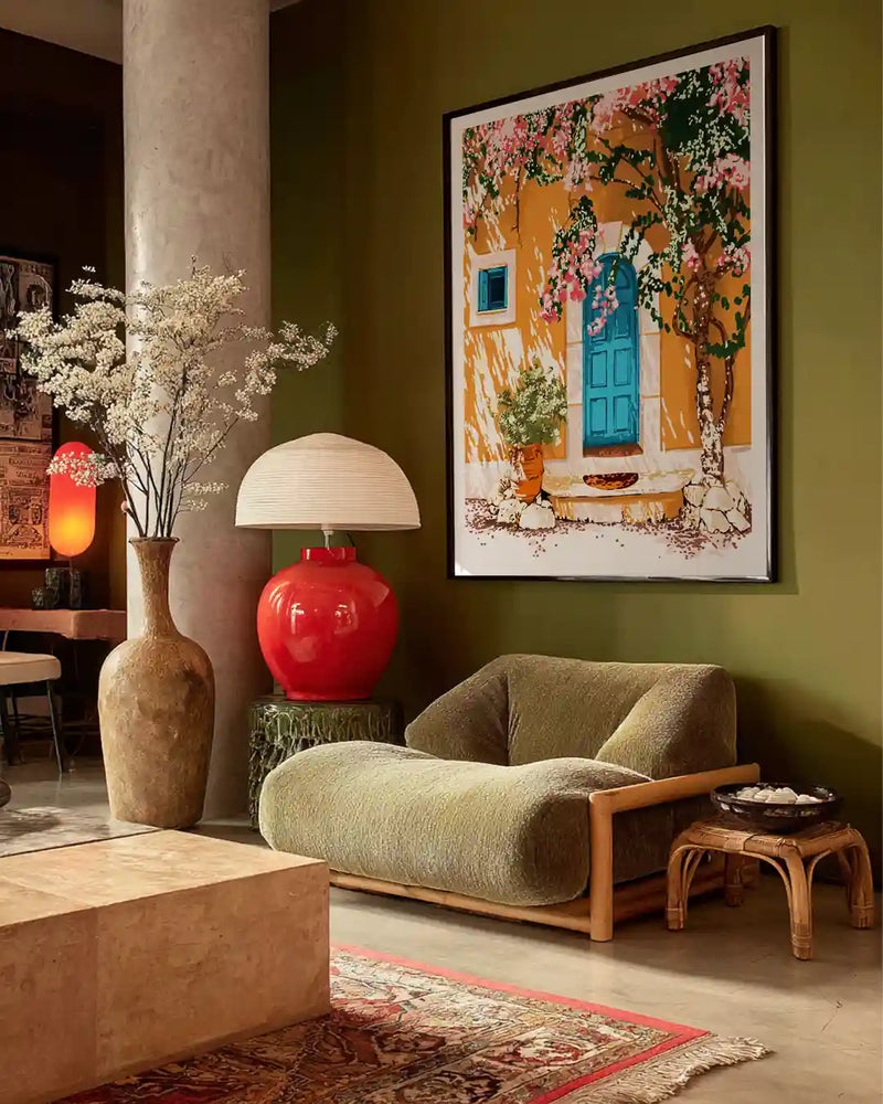 A cozy boho-style living room featuring a framed Mediterranean wall art print with a yellow house, blue door, and blooming bougainvillea.The artwork is styled above a modern lounge chair against an olive green wall, creating a warm, artistic interior.Earthy decor elements like a red table lamp, ceramic vase with dried flowers, and a patterned rug enhance the aesthetic.Perfect inspiration for bohemian home decor, wall art styling, and contemporary living room interiors.