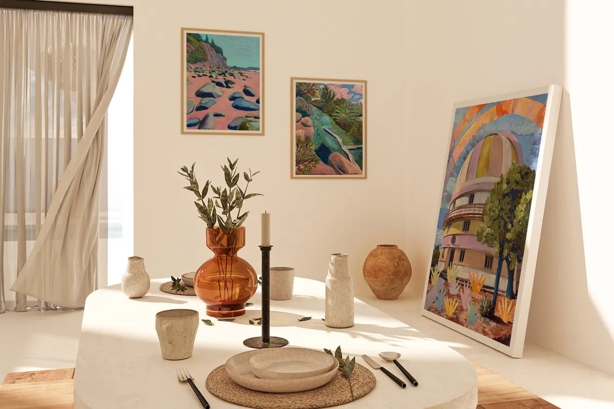 Sunlit dining corner with three colorful framed artworks, including one large frame leaning against the wall. Minimal table decor with ceramic vases, candles, and earthy-toned accents on a round table, soft curtains draped open beside a window.